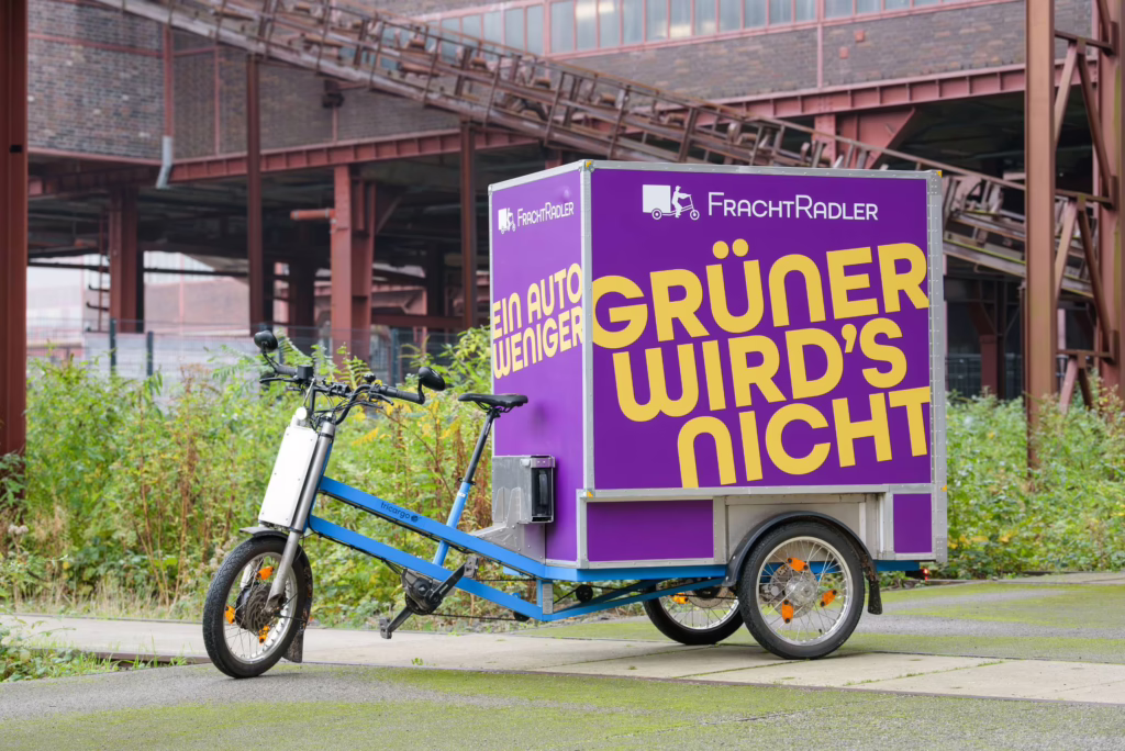 Ein Lastenrad von FrachtRadler. Das Rad wird von der Seite gezeigt, damit man einen Eindruck vom Fahrzeug bekommt. Es handelt sich um ein Dreirad mit großer Box hinten. Auf der Box steht neben dem FrachtRadler Logo "Grüner wird's nicht".