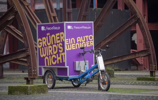 Ein Lastenrad von FrachtRadler. Das Rad wird von der Seite gezeigt, damit man einen Eindruck vom Fahrzeug bekommt. Es handelt sich um ein Dreirad mit großer Box hinten. Auf der Box steht neben dem FrachtRadler Logo "Grüner wird's nicht".